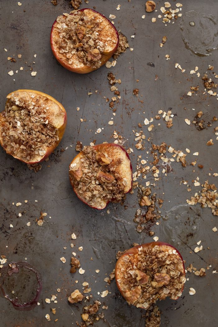 Apples cut in half on a baking sheet with walnuts, oats, and cinnamon sprinkled on top.