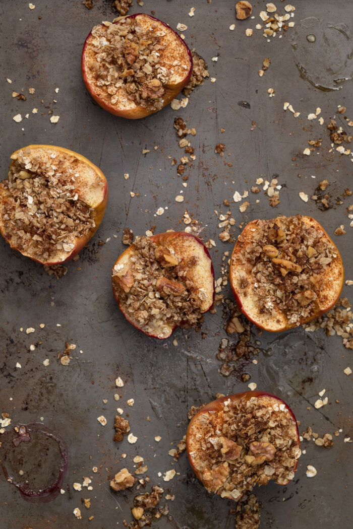 Apples cut in half on a baking sheet with walnuts, oats, and cinnamon sprinkled on top.