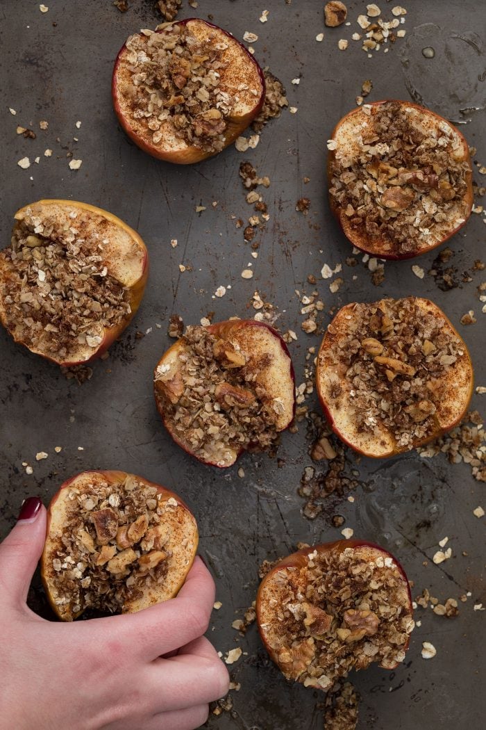 A hand reaching in to grab apples cut in half on a baking sheet with walnuts, oats, and cinnamon sprinkled on top.