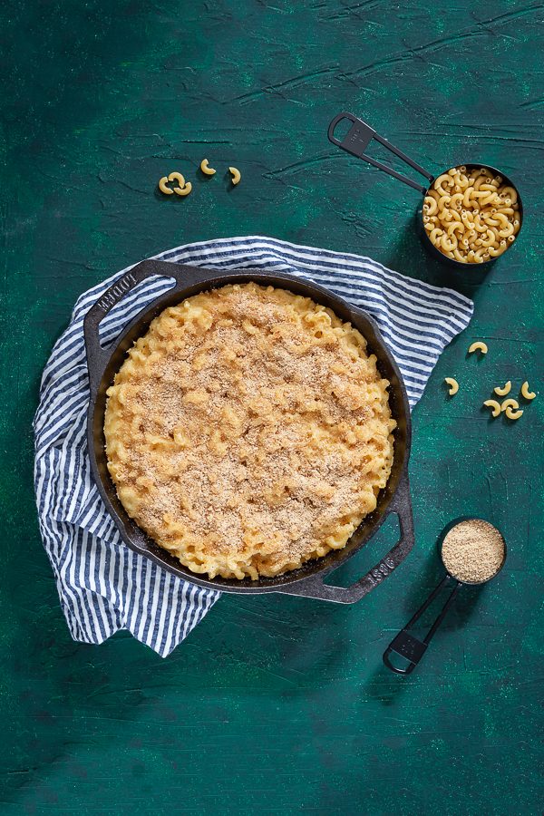 Macaroni and cheese baked in a cast iron skillet with elbow noodles and bread crumbs in measuring cups nearby. 