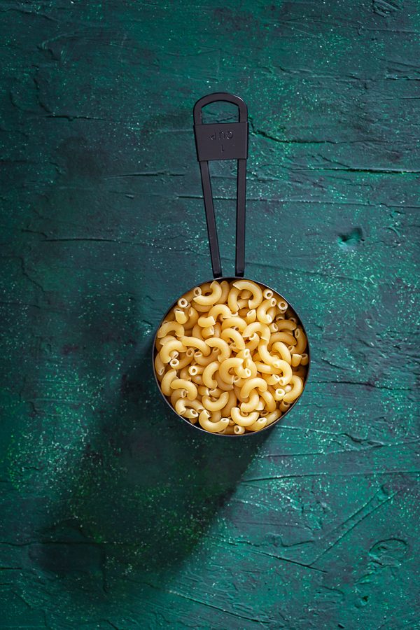 Black measuring cup on green background filled with yellow elbow noodles. 