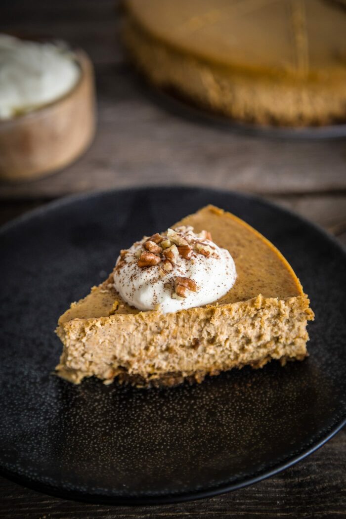 A slice of pumpkin spice cheesecake topped with whipped cream and pecans on a black plate and wood background.