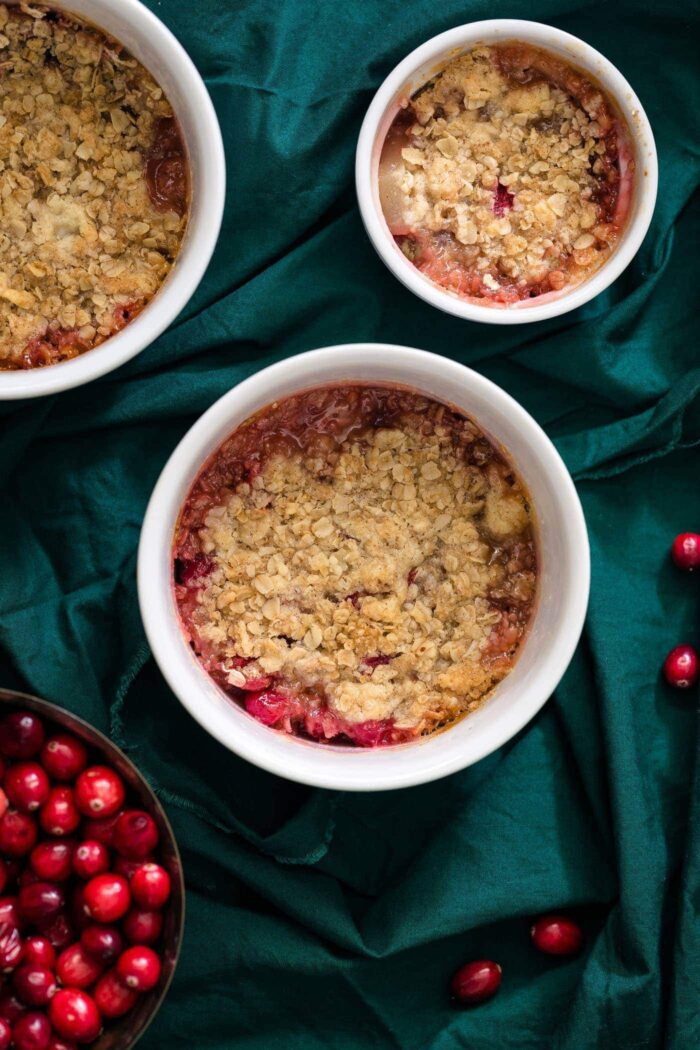 3 ramekins filled with cranberry and pear crisp topped with oats. A bowl of red cranberries nearby. 
